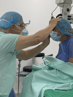 En un ambiente médico hay un paciente sentado con ropa quirúrgica y envuelto en mantas quirúrgicas, al frente de él hay un profesional de la salud revisando sus ojos.